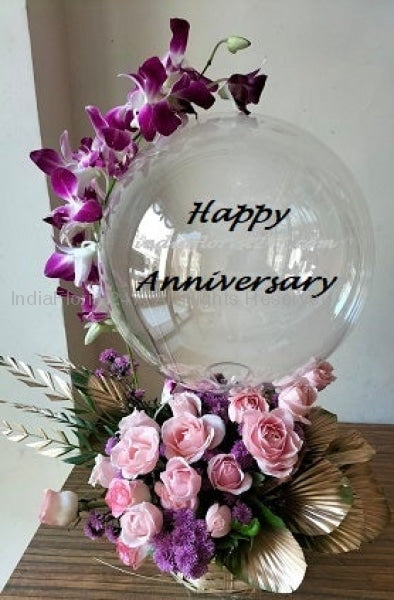 Print text with happy anniversary on clear balloon with orchid trailing on the balloon perched on top of pink roses with Gold colour leaves C-BFST