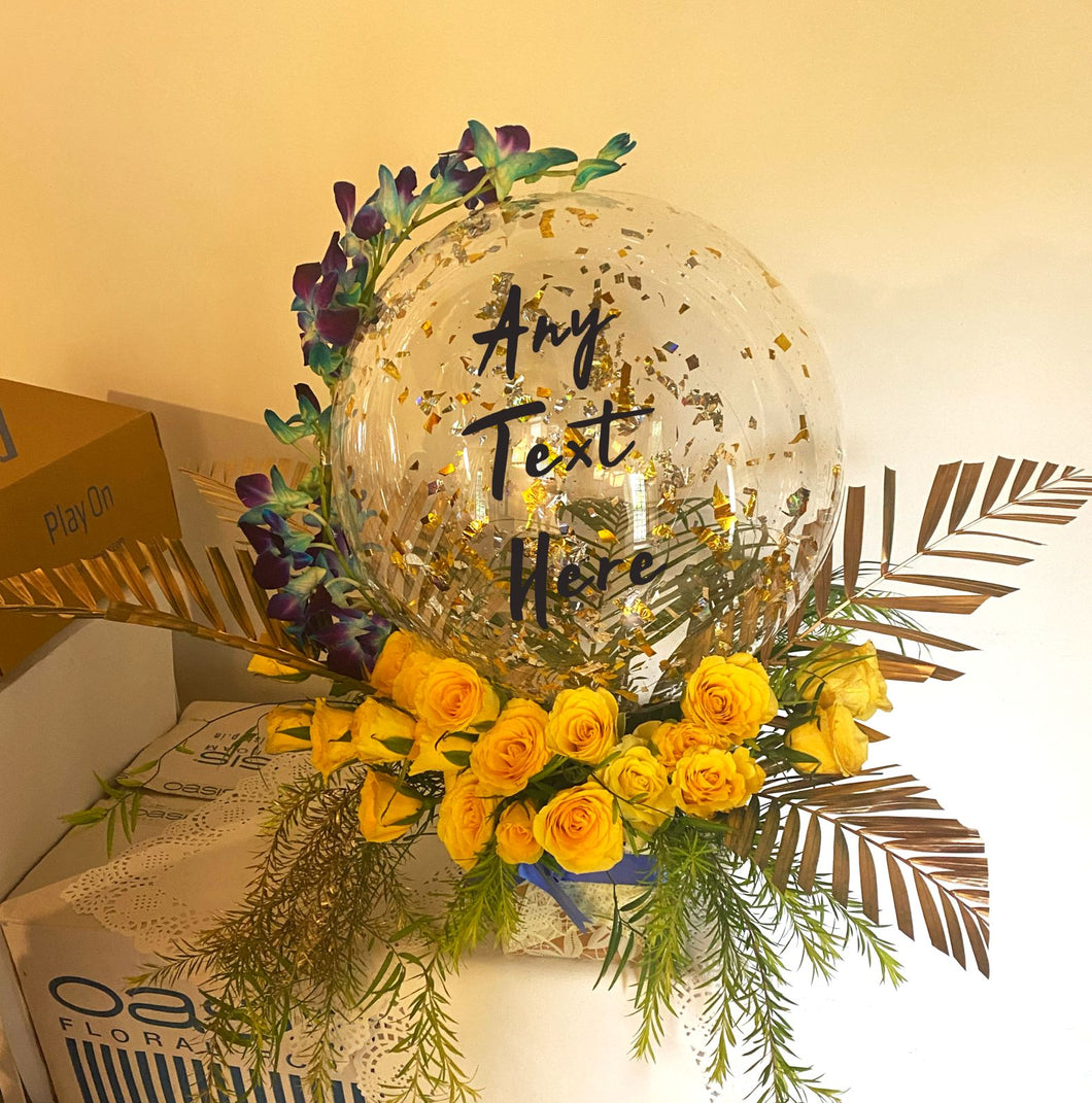 Print Any text and customise clear birthday balloon with orchid trailing on the balloon perched on top of yellow roses with Gold colour leaves C-BFST